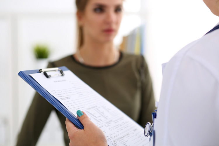 A doctor is holding a clipboard with a report on it and talking to a young woman. This illustrates the Medical Testing step that every donor participates in when preparing to donate stem cells or bone marrow with Gift of Life Marrow Registry.